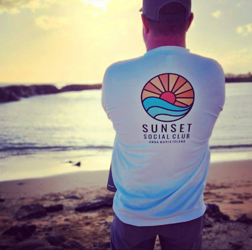 Person wearing a white long-sleeve Sunset Social Club shirt by Gnarly Mangrove Designs on Anna Maria Island during sunset at the beach.